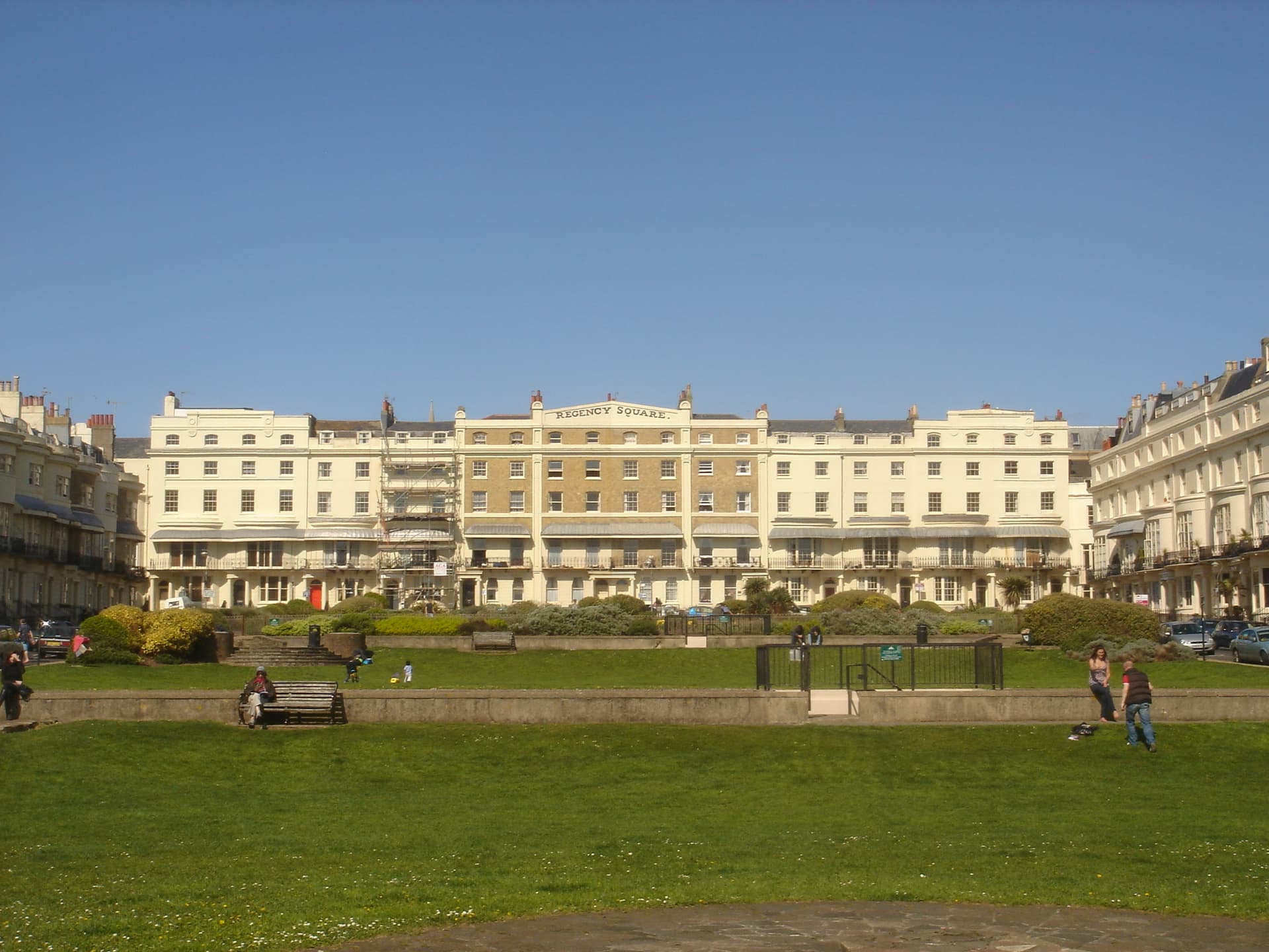Cream Regency terraces facing a grassy garden square under a clear blue sky, with people sitting on benches