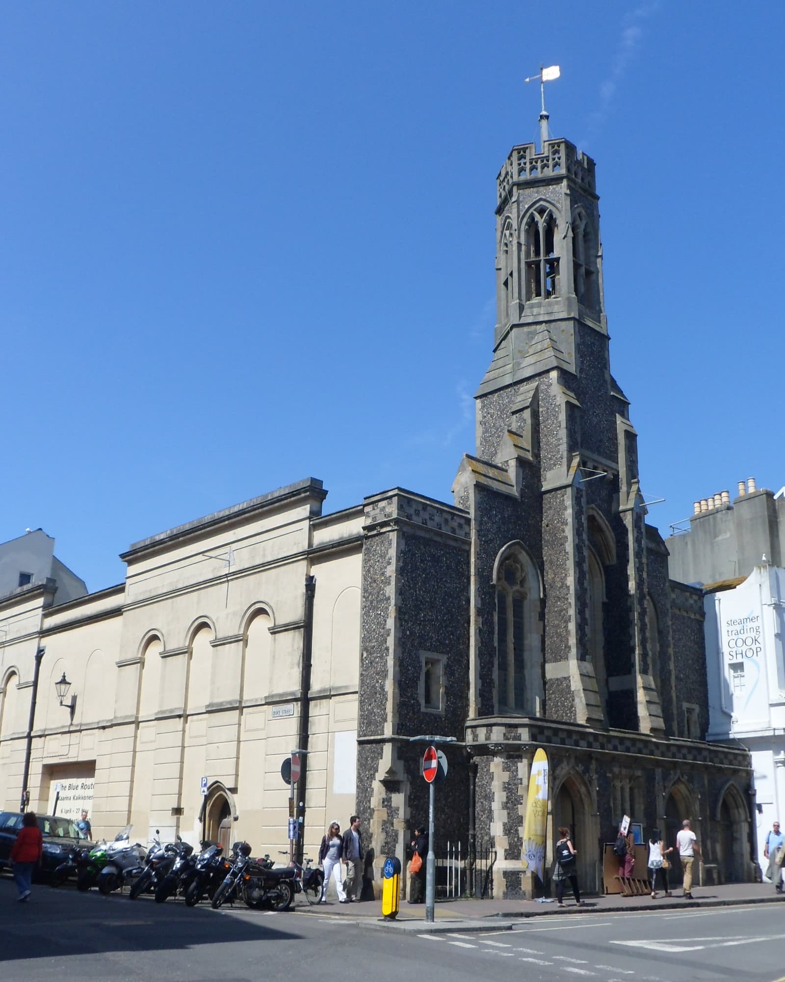 Flint-faced Gothic church tower and cream stucco side wall on a Brighton street corner under blue sky