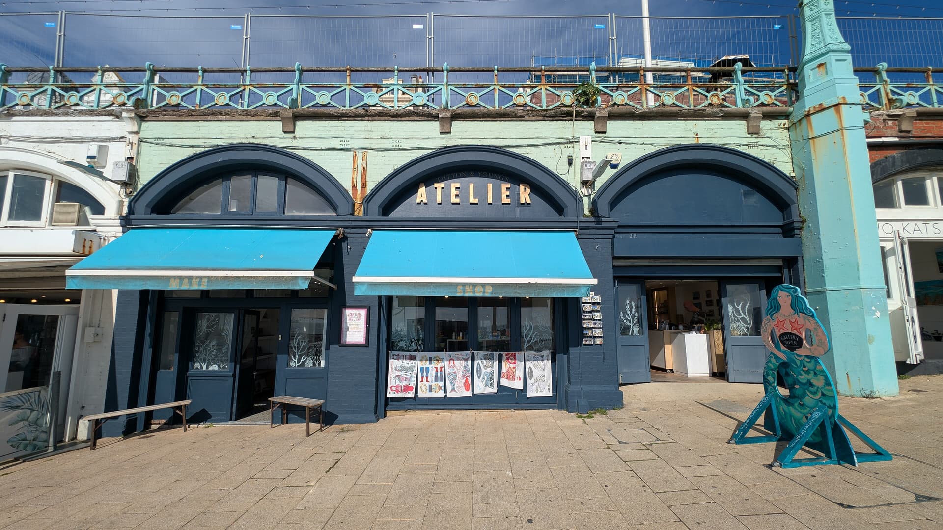Navy-painted seafront arches on Brighton's Kings Road with turquoise awnings, a mermaid cut-out and ornate cast-iron railings above