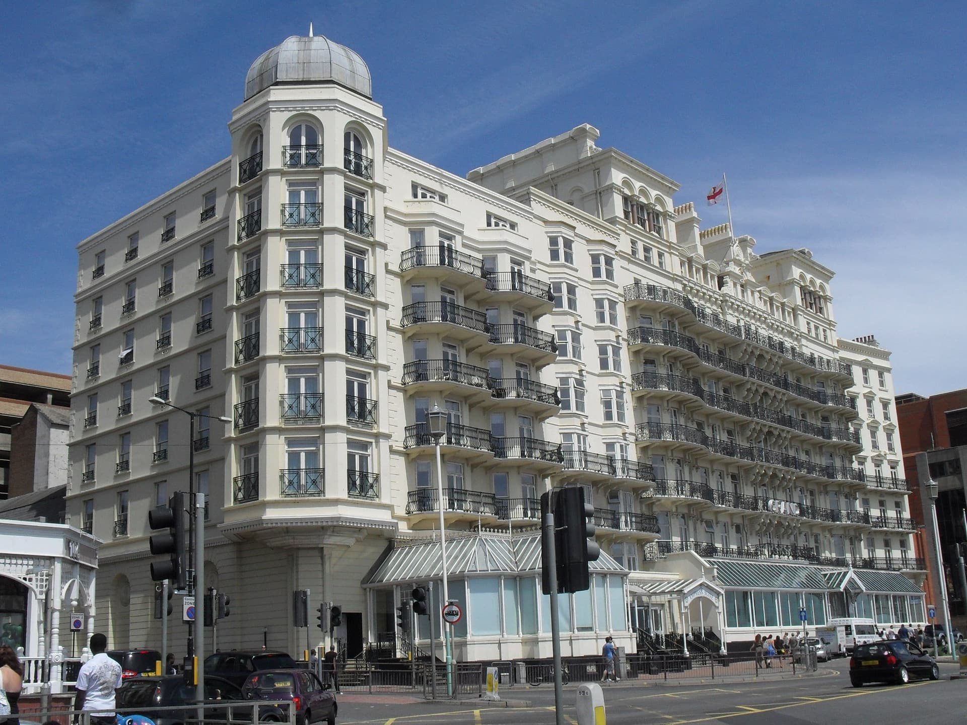 Cream Victorian seafront hotel with curved iron balconies, corner dome and glazed conservatory beneath a bright blue sky